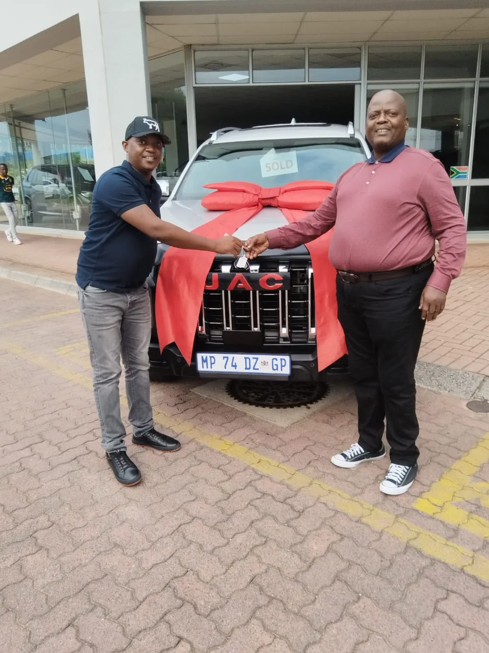 A smiling customer shakes hands with a salesperson as he receives the keys to his new silver JAC, decorated with a large red bow and a "Sold" sign.