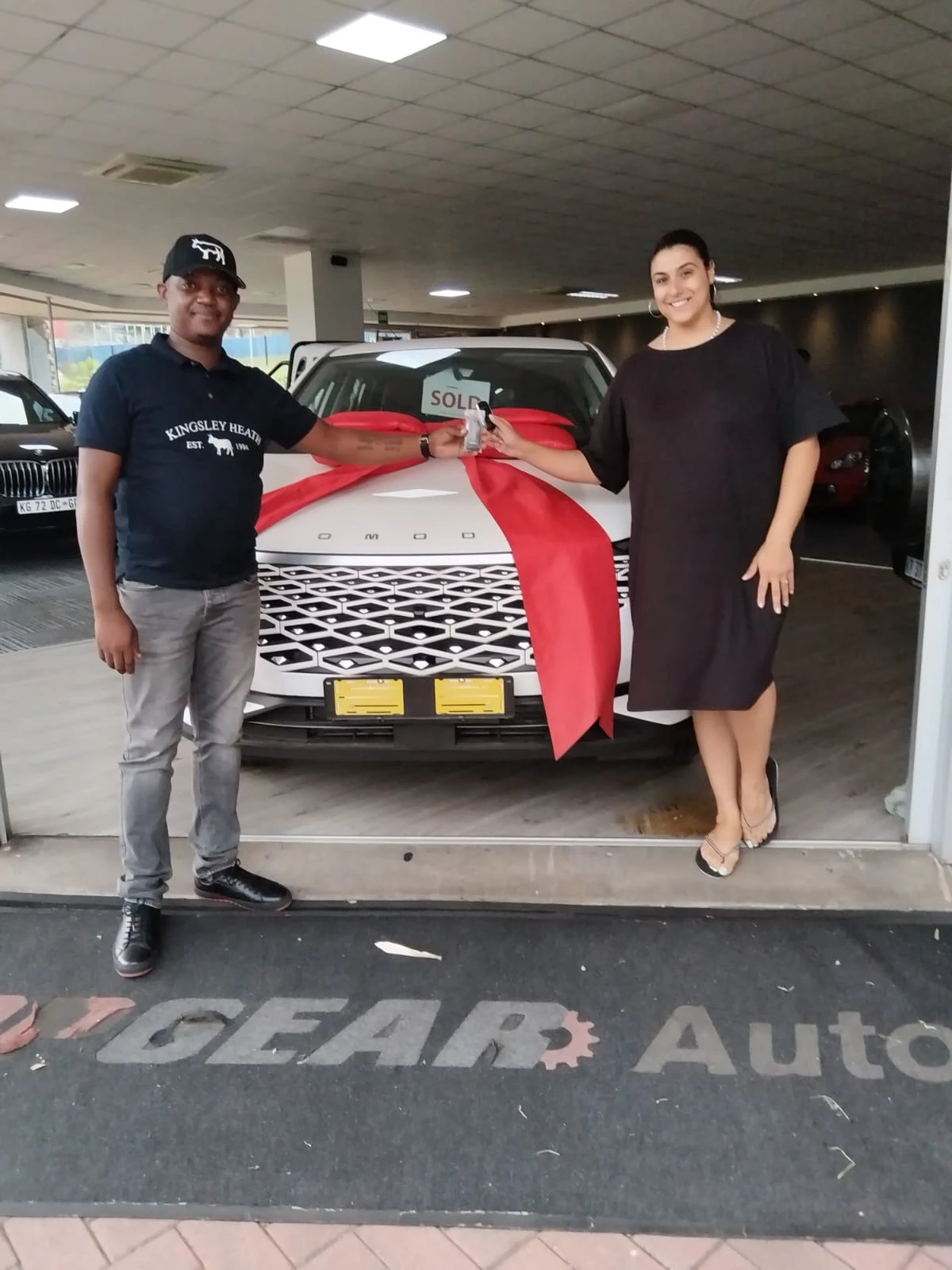 A salesperson hands a happy customer the keys to her new white Omoda SUV, wrapped in a big red bow, at the entrance of the TopGear Auto showroom.